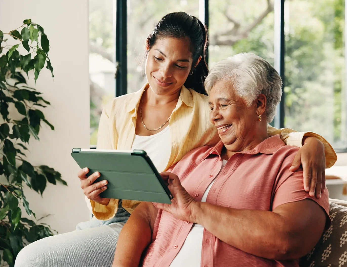 A woman sits with her arm around her mother on a sofa in a bright room, the two of them looking at a tablet together.